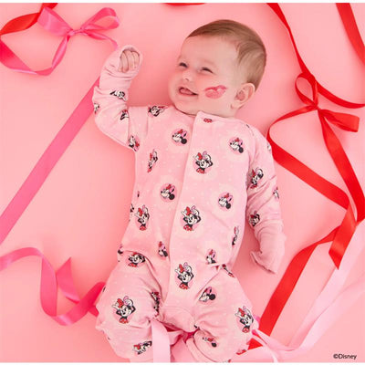Baby wearing a pink Minnie Mouse-themed coverall with red ribbons on a pink background, smiling with a kiss mark on the cheek.