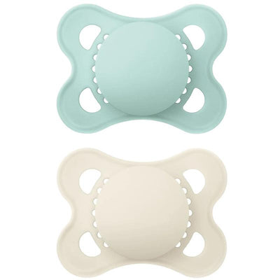 Two assorted Mam matte non-deco pacifiers for infants, one in light blue and the other in cream, featuring round nipples and a soft, curved shield.