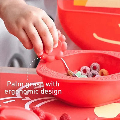 Toddler’s hand using a red ergonomic spoon to scoop colorful cereal from a matching bowl in a mealtime set.