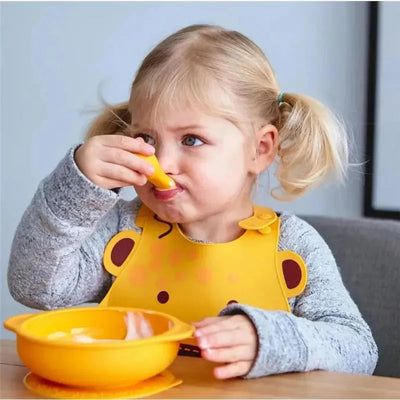 Toddler using a yellow spoon from the Marcus & Marcus self-feeding set while sitting at a table with a matching bowl.