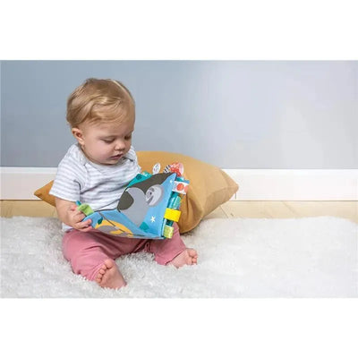 Baby sitting on a rug, exploring the Mary Meyer Taggies Molasses Sloth Book with colorful tags and a sloth design.
