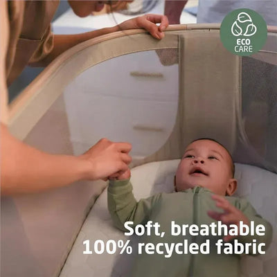 Maxi-Cosi Iora bedside bassinet in Classic Green features a baby reaching for a hand, with text highlighting soft, breathable, eco-friendly fabric.
