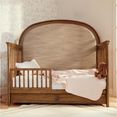 Maxi Cosi Quiet Luxury 6-in-1 Convertible Crib in natural wood finish, featuring a toddler bed configuration with a pink blanket and plush toy.