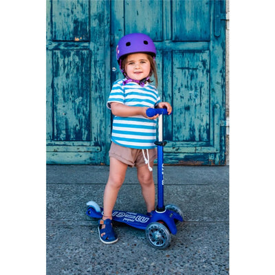 Child smiling while holding a Micro Kickboard Mini LED Scooter in blue and white, wearing a purple helmet, against a rustic blue door.