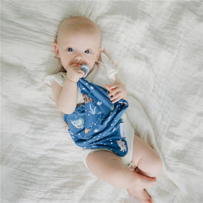 Baby holding a blue Finding Nemo lovey security blanket while sucking a pacifier, lying on a white blanket.