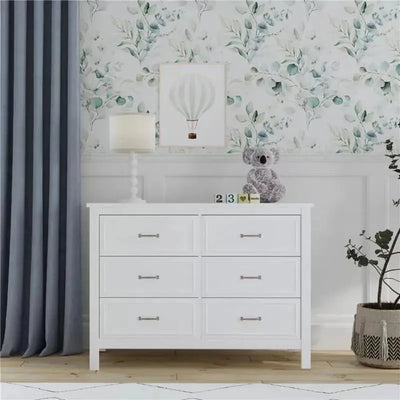 White six-drawer dresser with silver handles, beside a lamp, decorative items, and blue curtains against a floral wall.