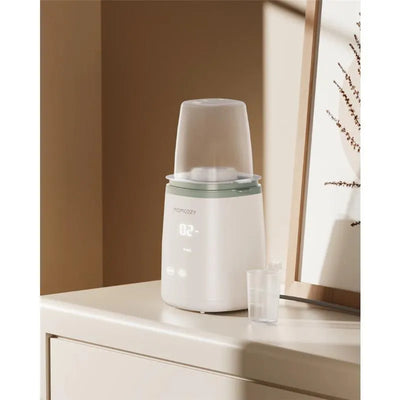 Momcozy 6-in-1 Fast Baby Bottle Warmer on a bedside table, featuring a digital display and measuring cup beside it.