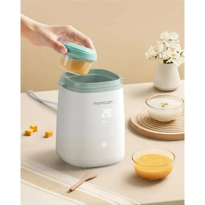 Momcozy 6-in-1 Fast Baby Bottle Warmer with a hand placing a container inside, and bowls of food on a light surface.