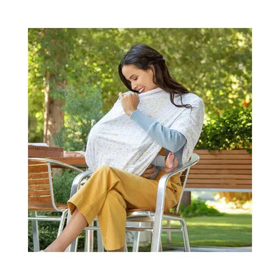 A woman sits outdoors in a chair, using the Munchkin Antimicrobial 5-In-1 Cover, gently smiling as she nurses a baby.
