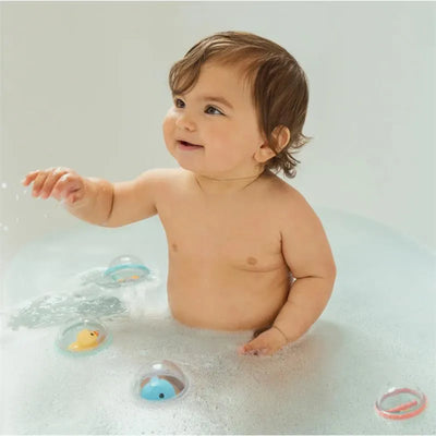 A smiling baby in a bath with bubbles, reaching for colorful floating toys inside clear plastic bubbles.