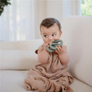 Baby sitting on a couch wearing a natural-colored, organic cotton wearable blanket, holding a green silicone teething toy.