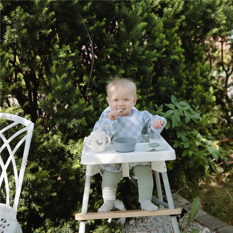 Toddler sitting in a high chair, wearing a blue checkered bib, with a silicone training cup and bowl on the tray.