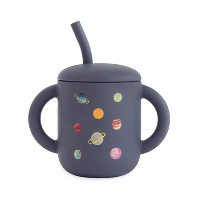 Mushie silicone training cup with a dark blue body, featuring colorful planet designs and a matching straw.
