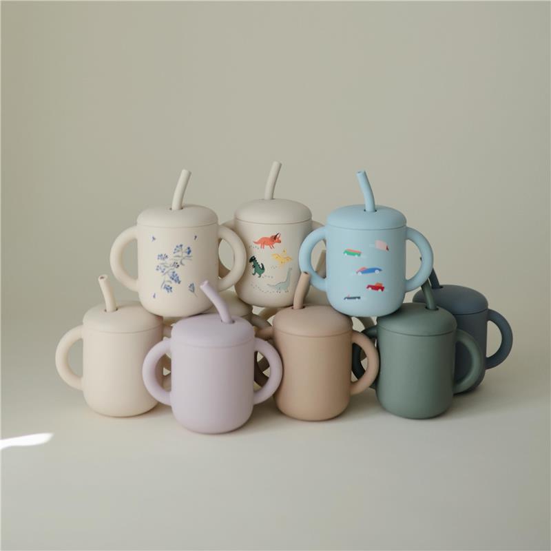 Mushie silicone training cups in assorted colors with playful designs, featuring handles and built-in straws for toddlers.