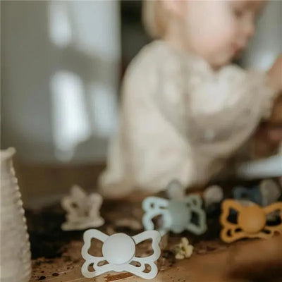 Butterfly-shaped silicone pacifiers in gray and pastel colors, designed for infants aged 6 to 18 months.