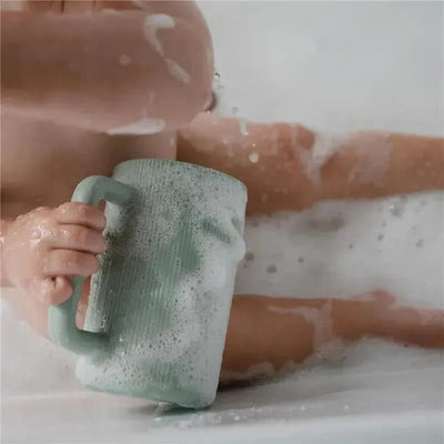 Mushie Bath Rinse Cup in Cambridge Blue held by a child with bubbly water in a bathtub setting.