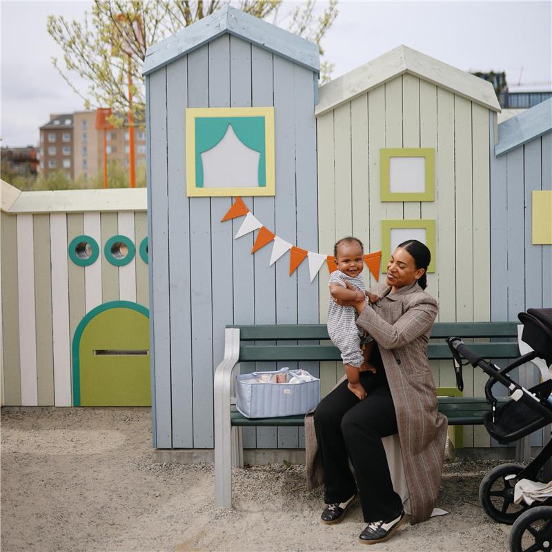 Mother holding a smiling baby near a blue floral diaper caddy on a park bench, with colorful playhouse structures in the background.