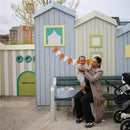 Mother holding a smiling baby near a blue floral diaper caddy on a park bench, with colorful playhouse structures in the background.