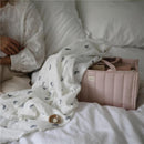 Mushie Baby Diaper Caddy in blush color sits on a bed, partially covered by a patterned blanket, with a hand resting nearby.