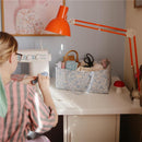 A person in a striped shirt stitches with a sewing machine at a desk, next to a pastel floral diaper caddy filled with fabric.