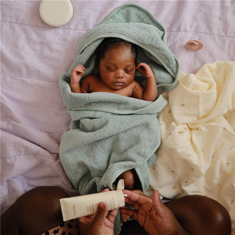 Hooded towel in sea mist color wrapped around a sleeping baby, with lotion in hand and soft fabrics nearby.
