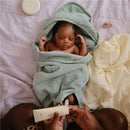 Hooded towel in sea mist color wrapped around a sleeping baby, with lotion in hand and soft fabrics nearby.