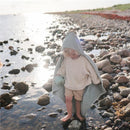 Baby wearing a sea mist organic cotton hooded towel, standing on rocky beach shore, holding a green toy.