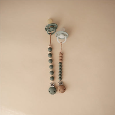 Mushie Eva pacifier clip in Dried Thyme features two pacifier holders with rounded beads in muted green tones and a wooden clip.