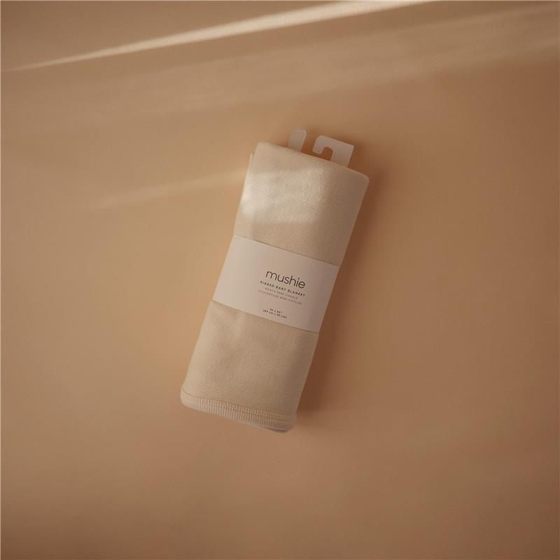 Mushie Extra Soft Baby Blanket in ivory, neatly rolled and displayed with a white label on a beige background.