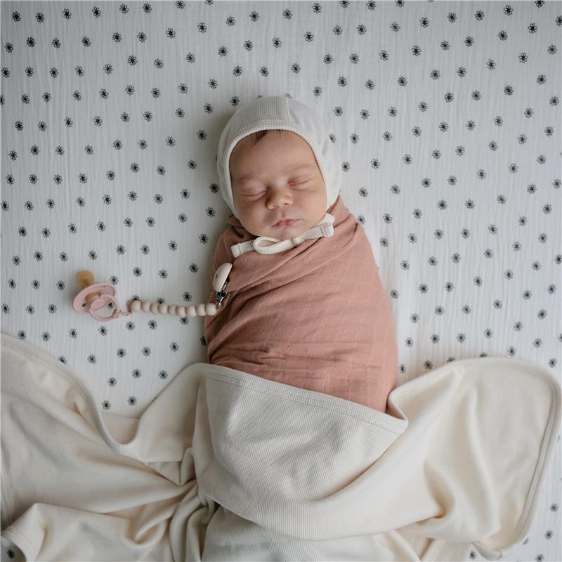 Ivory Mushie Extra Soft Baby Blanket drapes softly around a sleeping baby, who wears a beige swaddle and a knitted cap.