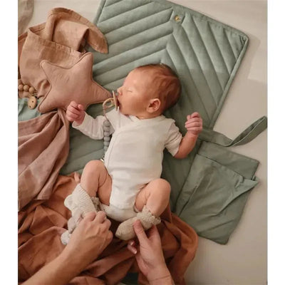 A baby wearing a white onesie and knitted socks, sucking on a pacifier, rests on a green quilted mat, surrounded by soft fabric and a star-shaped cushion.