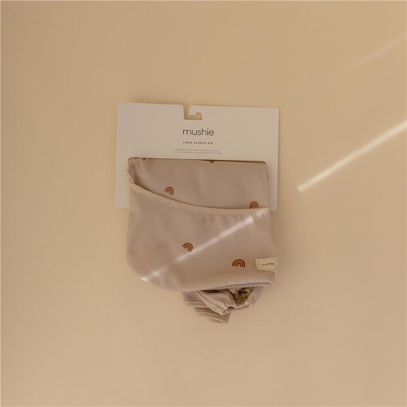 Mushie long sleeve baby bib in beige with rainbow patterns, featuring adjustable fit and water-resistant material.