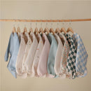 Water-resistant baby bibs in various colors and patterns, including dinos and floral designs, hung on wooden hangers.
