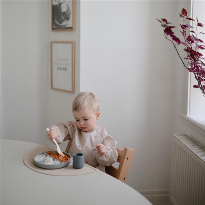 Toddler wearing a long sleeve water-resistant bib, eating from a plate with rice and food, at a light-filled dining area.