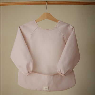 Mushie Long Sleeve Bib in blush color, featuring elasticized cuffs and a pocket at the bottom for catching spills.