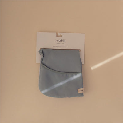 Mushie Long Sleeve Bib in Tradewinds color, displayed on a hanging tag against a beige background.
