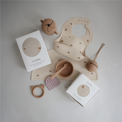 Silicone baby bib featuring rainbow designs with additional Mushie baby products including dishes and a rattle.