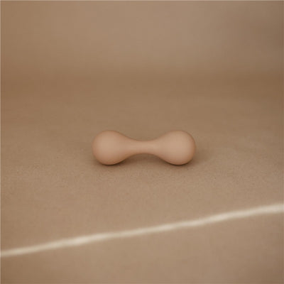 Mushie silicone baby rattle toy in a neutral color, featuring an hourglass shape, resting on a textured surface.