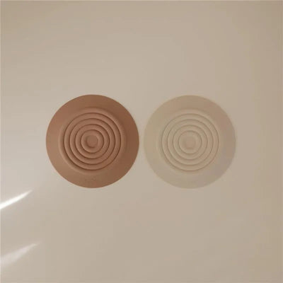 Mushie silicone drain stoppers in natural and shifting sand colors, featuring a concentric circular design.