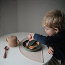 Child using a pink fork to eat orange segments from a green silicone divided plate on a beige silicone placemat.