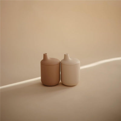 Mushie Silicone Sippy Cups in natural tones, featuring a rounded design and small spout, positioned on a soft surface.