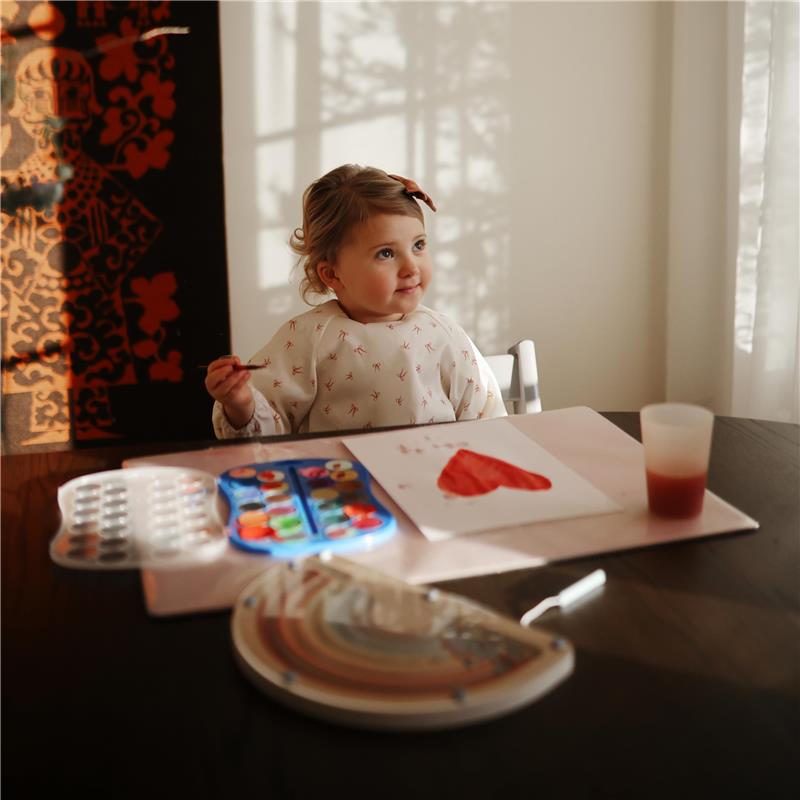 A young child wearing a bow and a long sleeve baby bib is seated at a table, holding a paintbrush and smiling, with a painted heart on paper and art supplies nearby.