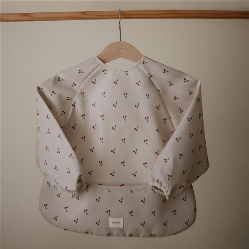 Water-resistant long sleeve baby bib with cherries pattern, featuring adjustable fit and elastic cuffs, by Mushie.