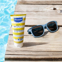 Mustela Mineral Sunscreen Lotion SPF 50, 3.38 fl oz, placed on wooden surface beside blue sunglasses, with swimming pool in background.