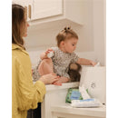 Toddler sitting on a countertop, reaching for Mustela Compostable Cleansing Water Wipes beside a warmers.