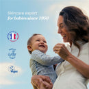Mustela Cradle Cap Cream displayed with a mother holding her smiling baby outdoors; text highlights skincare expertise since 1950.
