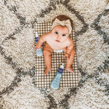 A baby sits on a checkered mat surrounded by two bottles of Mustela Hair Styler & Skin Freshener, featuring light blue packaging.