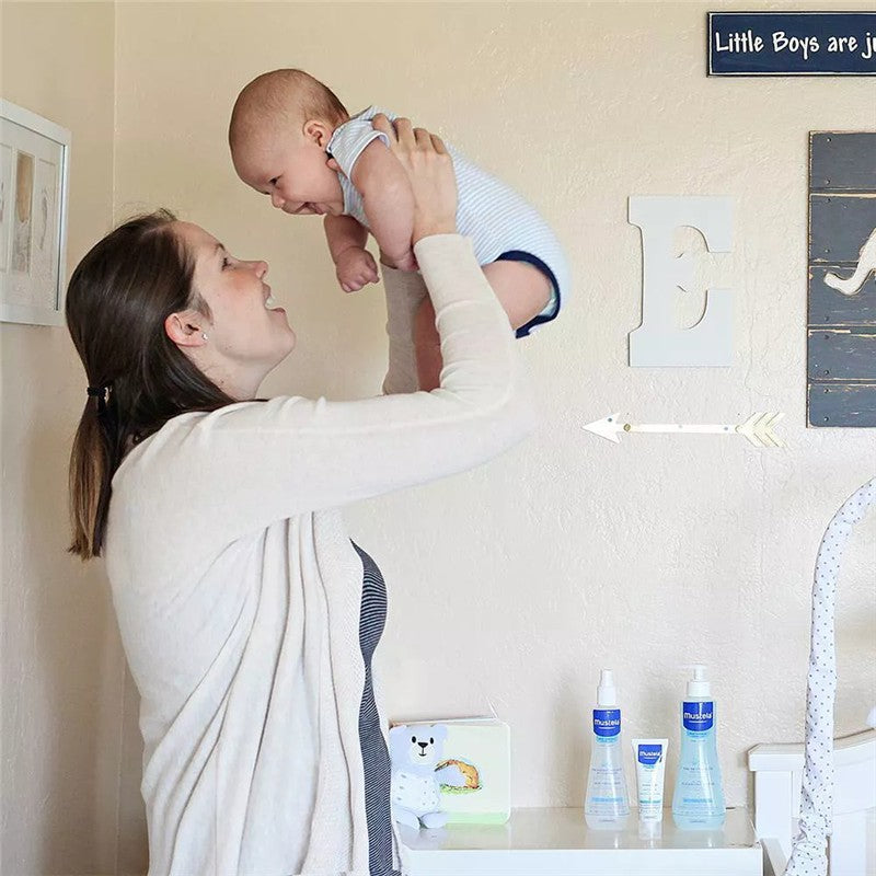 A mother joyfully lifts her baby in a nursery, with Mustela hair styler and skin freshener products visible on the shelf.
