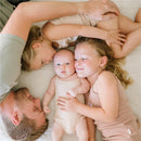 A father and two young sisters cuddle on a bed, with one sister holding the newborn baby who wears a light onesie.