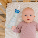 Mustela No-Rinse Cleansing Water bottle next to a baby in a crib, featuring a light pink outfit and a neutral background.